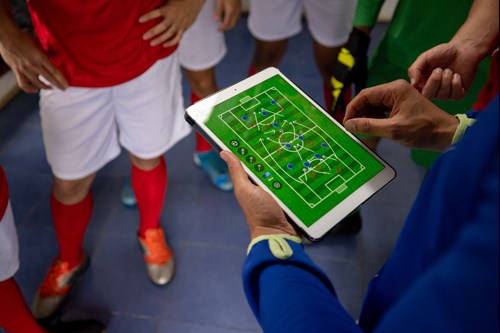 A coach using a tablet to show a digital soccer tactics board to players wearing red jerseys and white shorts in a locker room.
