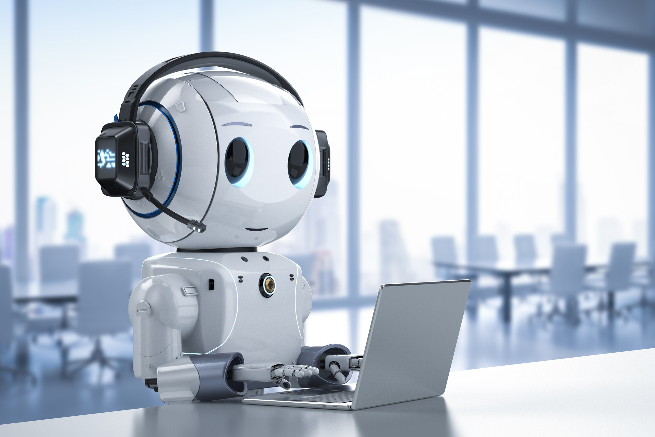 A friendly-looking robot with a round head, wearing large headphones with a microphone, sitting at a desk and using a laptop in a modern, well-lit office environment.