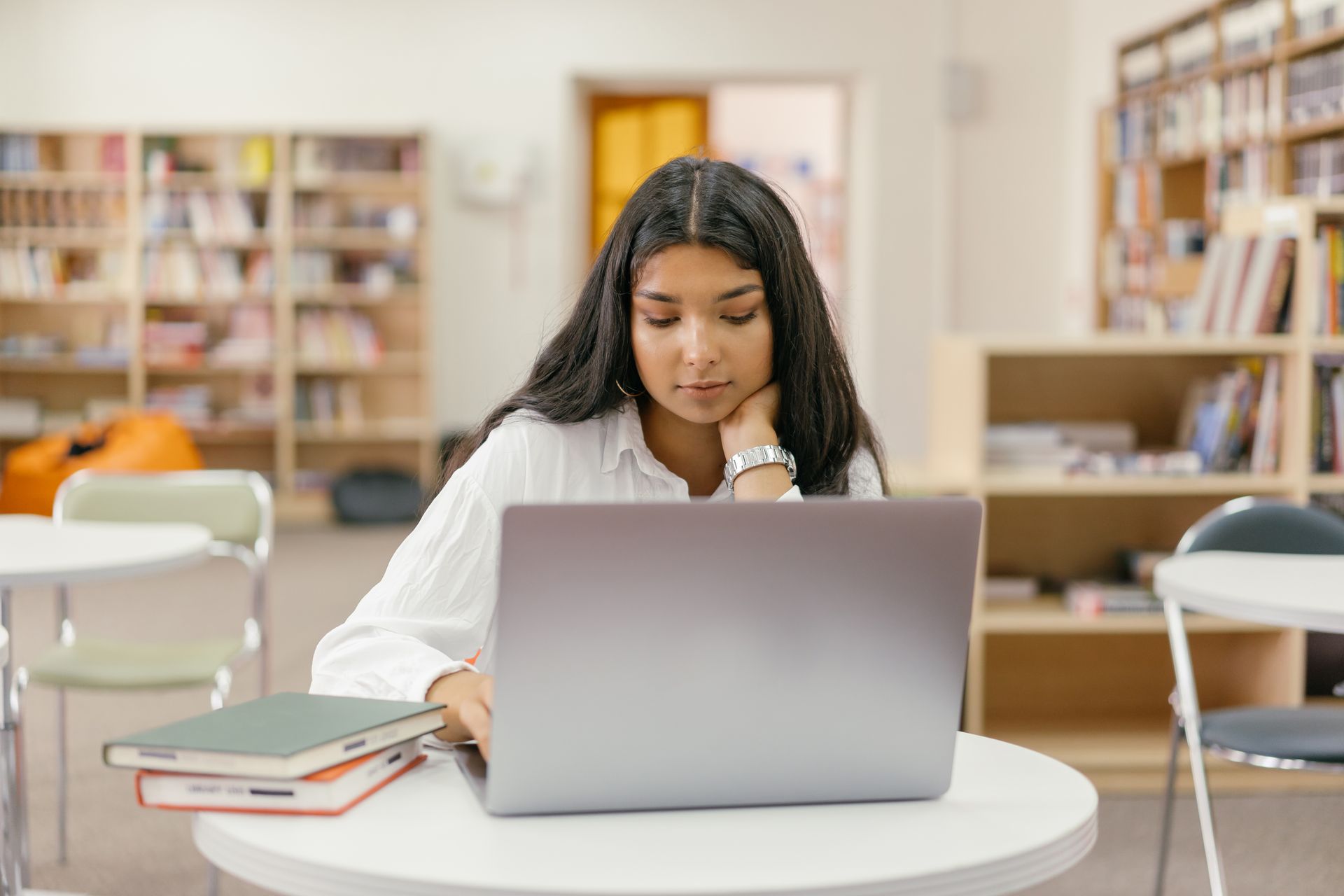 A person sitting at a table in a library, using a laptop, with a stack of books nearby.