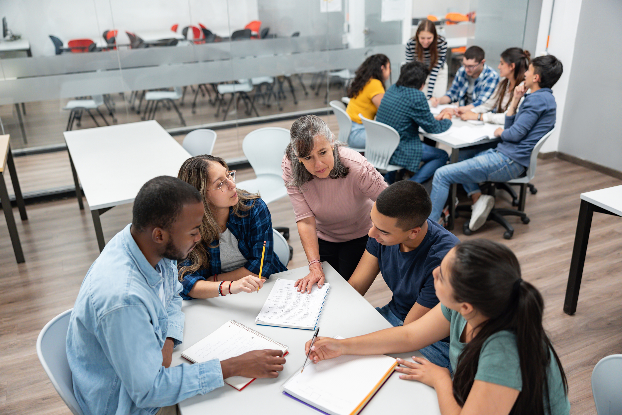 A teacher is interacting with a diverse group of students sitting around a table, engaged in a study session, in a modern classroom with glass walls and additional groups in the background.