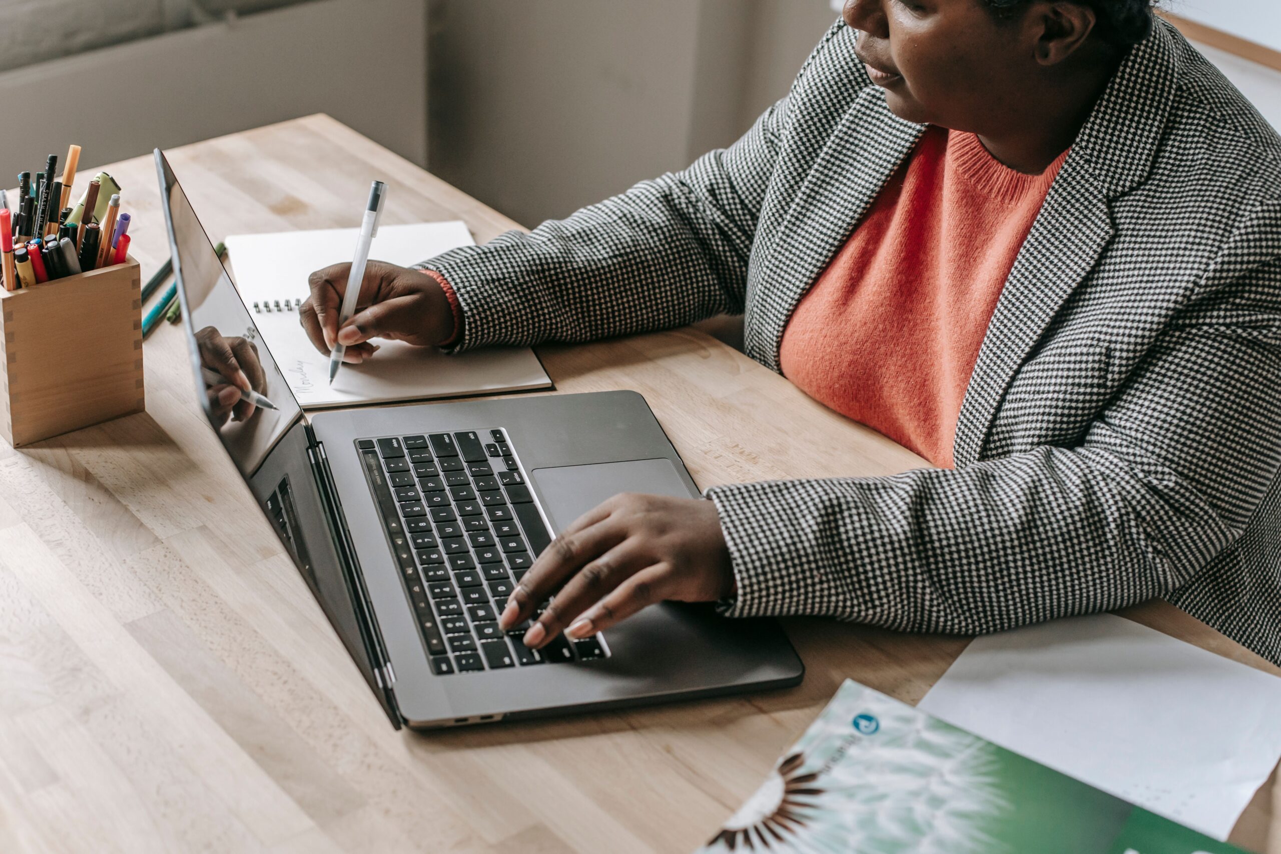 Professional woman in a formal outfit working on a laptop and writing notes at a desk.